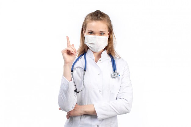 A front view young female doctor in white medical suit with stethoscope wearing white protective mask posing on the white