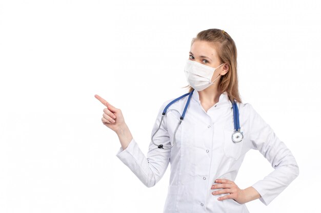 A front view young female doctor in white medical suit with stethoscope wearing white protective mask posing on the white