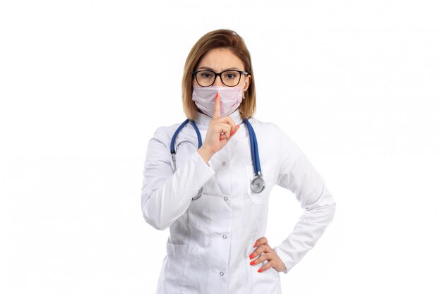 A front view young female doctor in white medical suit with stethoscope wearing white protective mask showing silence sign on the white