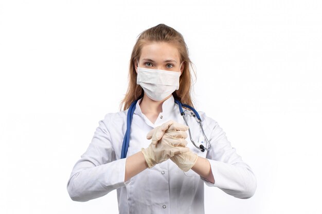 Вид спереди молодая женщина-врач в белом медицинском костюме со стетоскопом в белой защитной маске в перчатках на белом фоне
