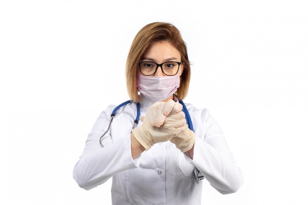 A front view young female doctor in white medical suit with stethoscope wearing white protective mask wearing gloves on the white