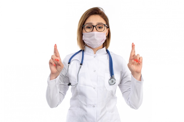 A front view young female doctor in white medical suit with stethoscope wearing white protective mask on the white