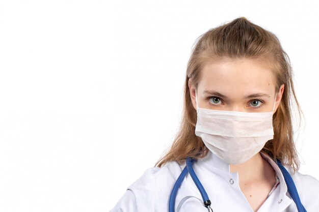 A front view young female doctor in white medical suit with stethoscope wearing white protective mask on the white