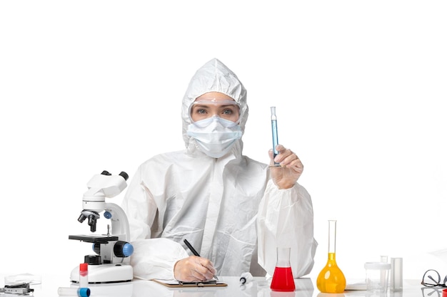 Вид спереди молодая женщина-врач в белом защитном костюме с маской из-за covid, держащая синий раствор на светло-белом фоне, всплеск пандемического вируса covid
