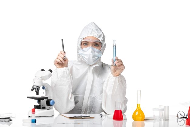 Front view young female doctor in white protective suit with mask due to covid holding blue solution on the white background splash pandemic covid- virus