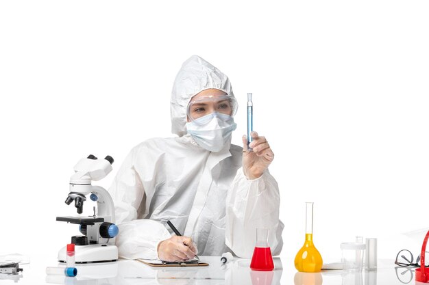 Вид спереди молодая женщина-врач в белом защитном костюме с маской из-за covid, держащая синий раствор на белом столе, всплеск пандемического вируса covid-
