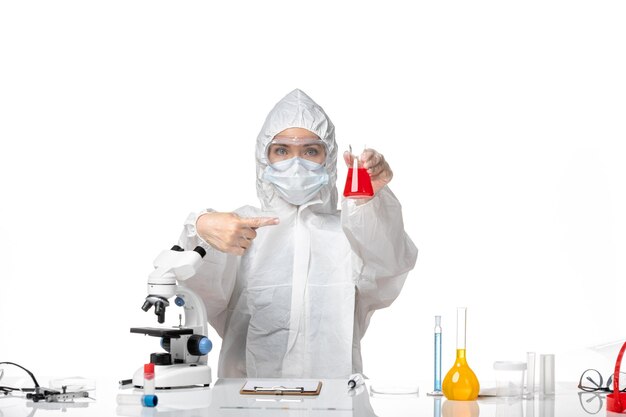 白い背景のウイルスパンデミックスプラッシュcovidに赤いソリューションを保持しているcovidによるマスクと白い防護服の若い女性医師の正面図