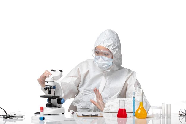 Front view young female doctor in white protective suit with mask due to covid working with microscope on light white background virus pandemic splash covid-