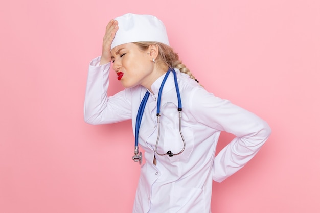 Вид спереди молодая женщина-врач в белом костюме с синим стетоскопом с головной болью на розовой космической женской работе
