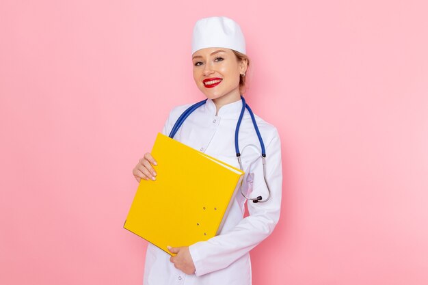 Front view young female doctor in white suit with blue stethoscope holding yellow files on the pink space  female work