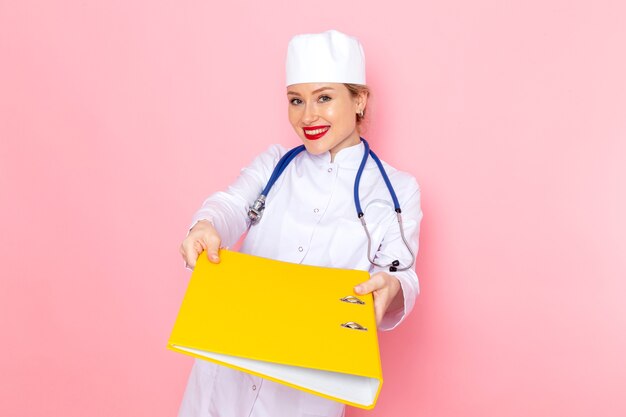 Front view young female doctor in white suit with blue stethoscope holding yellow files smiling on the pink space  female work