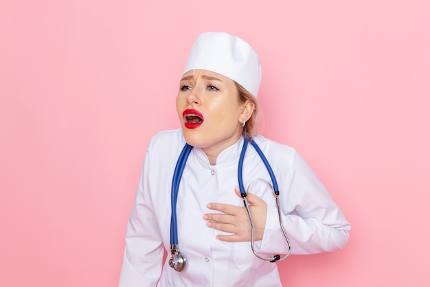 Giovane medico femminile di vista frontale in vestito bianco con lo stetoscopio blu che posa avendo dolore di cuore sul lavoro femminile dello spazio rosa