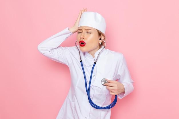 파란색 청진 기 핑크 공간 여성 작업에 그녀의 머리를 잡고 포즈와 흰색 정장에 전면보기 젊은 여성 의사