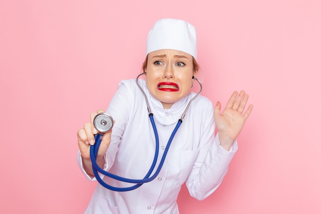 Front view young female doctor in white suit with blue stethoscope posing and measuring on the pink space  female work