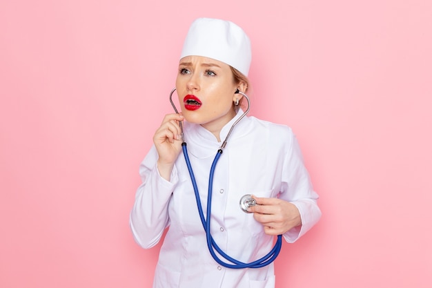 Front view young female doctor in white suit with blue stethoscope posing and measuring on the pink space  female work