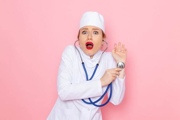 Front view young female doctor in white suit with blue stethoscope posing measuring with surprised expression on the pink space medicine medical hospital