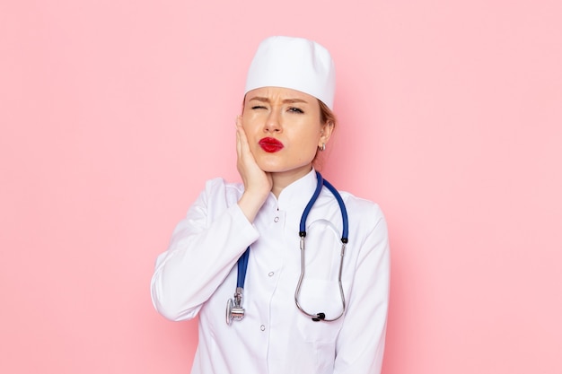 Medico femminile giovane di vista frontale in vestito bianco con lo stetoscopio blu che posa sulle emozioni della ragazza della donna della donna di lavoro dello spazio rosa