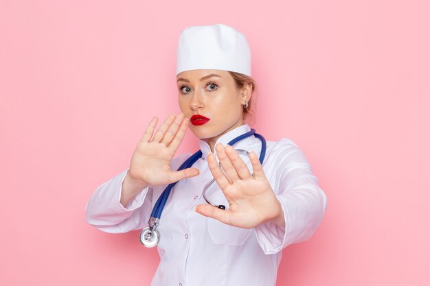 Front view young female doctor in white suit with blue stethoscope posing on the pink space job 