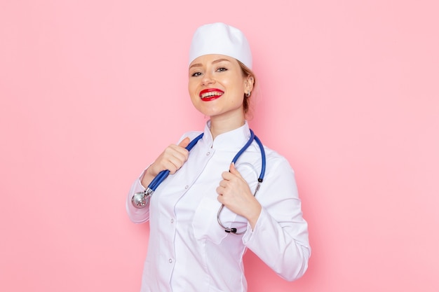Вид спереди молодая женщина-врач в белом костюме с синим стетоскопом позирует, улыбаясь на розовом космическом рабочем месте, врач медицинской больницы