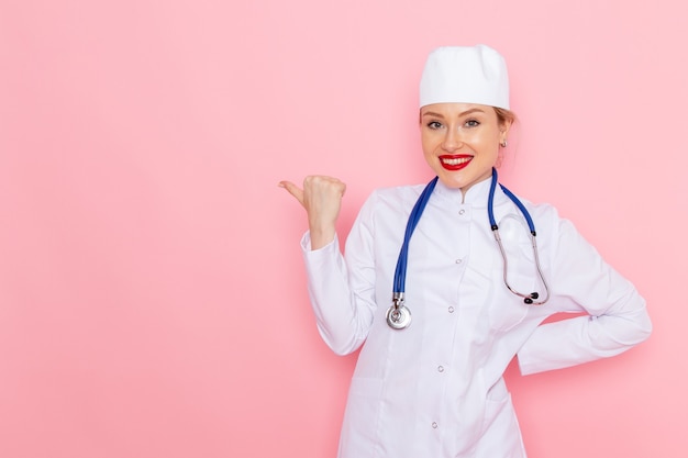 Вид спереди молодая женщина-врач в белом костюме с синим стетоскопом позирует и улыбается в розовой медицинской больнице космической медицины