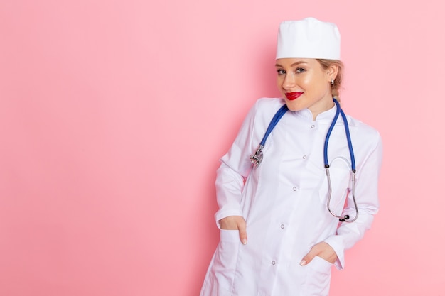 Front view young female doctor in white suit with blue stethoscope posing with smile on the pink space medicine medical hospital doctor female work