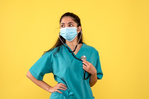 Вид спереди молодой женщины-врача с медицинской маской, держащей стетоскоп на желтой стене