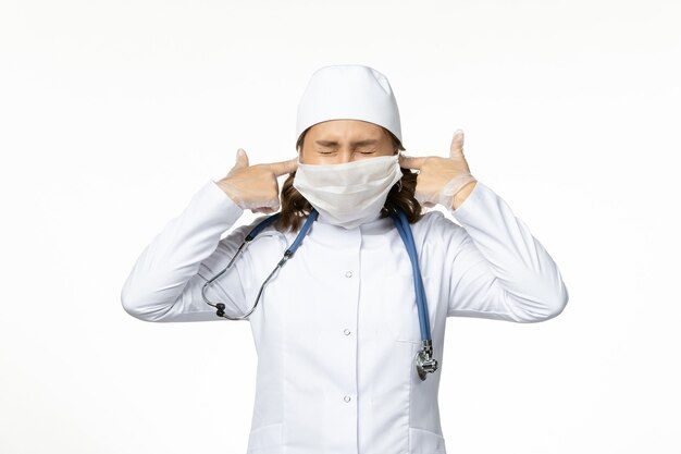 Front view young female doctor with protective mask due to coronavirus shutting her ears on white surface