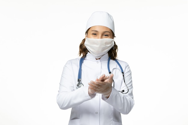 Front view young female doctor with protective mask due to coronavirus and smiling on the white surface