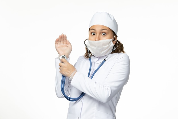 Front view young female doctor with protective mask due to coronavirus using stethoscope on white desk