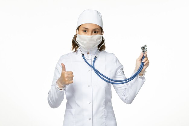 Front view young female doctor with sterile mask due to coronavirus holding stethoscope on white surface
