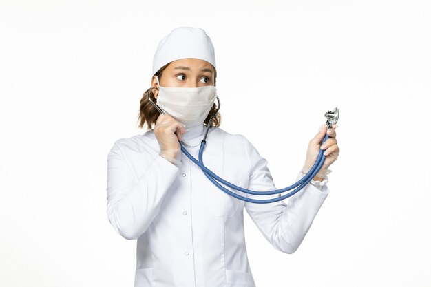 흰색 표면에 청진기를 사용하여 코로나 바이러스로 인해 멸균 마스크가있는 전면보기 젊은 여성 의사