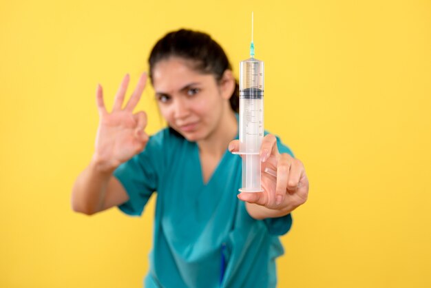 Front view of young female doctor with syringe making okey sign on yellow wall