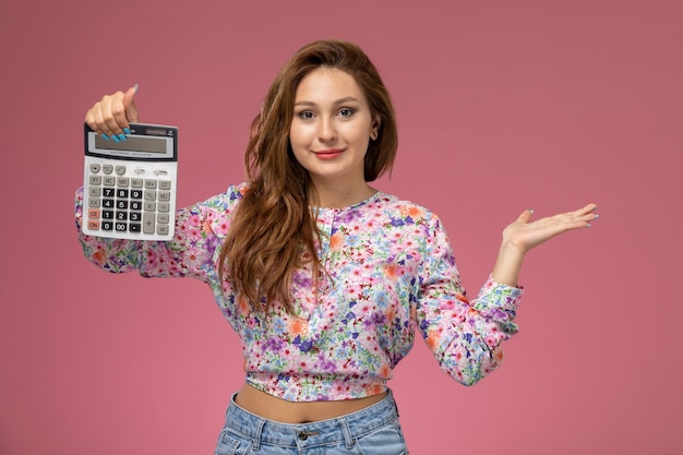 Front view young female in flower designed shirt and blue jeans holding a calculator and smiling on pink background 