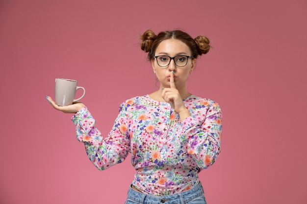 Free photo front view young female in flower designed shirt and blue jeans holding cup showing silence sign on the pink background