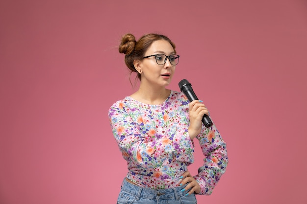 Front view young female in flower designed shirt and blue jeans holding mic trying to sing on the light background 