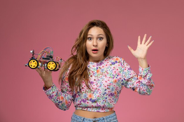 Front view young female in flower designed shirt and blue jeans holding a toy car on the pink background 