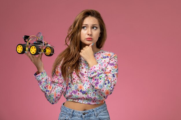 Front view young female in flower designed shirt and blue jeans holding toy car with thinking expression on the pink background