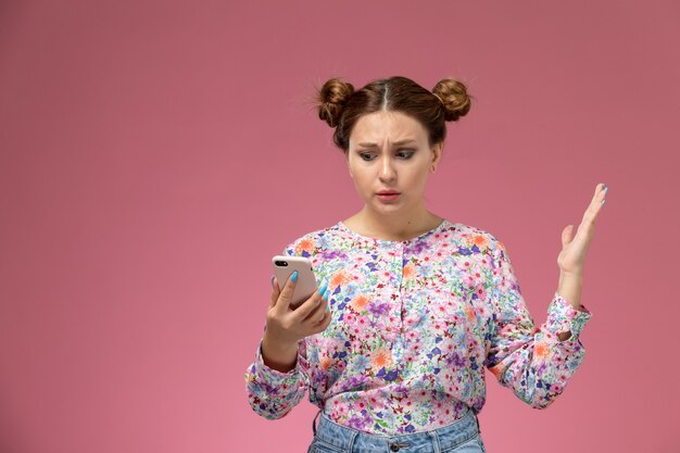 Front view young female in flower designed shirt and blue jeans holding and using a phone on the pink background 