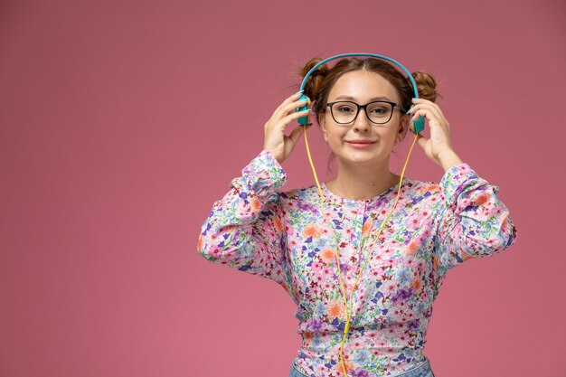 Front view young female in flower designed shirt and blue jeans listening to music with earphones on pink background