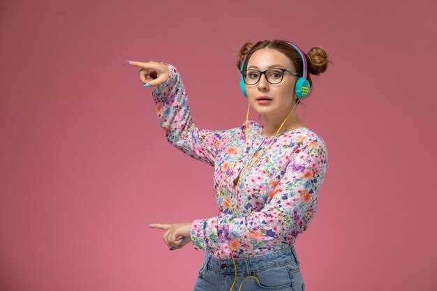 Вид спереди молодая женщина в рубашке с цветочным дизайном и синих джинсах, слушающая музыку в наушниках на розовом фоне