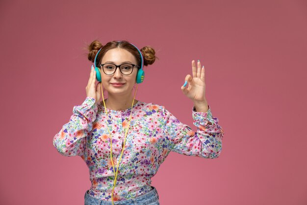 Front view young female in flower designed shirt and blue jeans listening to music with smile on the pink background