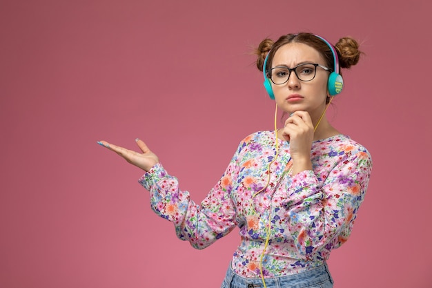 Вид спереди молодая женщина в рубашке с цветочным дизайном и синих джинсах слушает музыку с мысленным выражением на розовом фоне