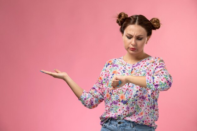 Front view young female in flower designed shirt and blue jeans looking at her wrist on the pink background 
