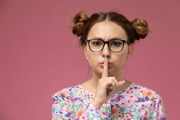 Vista frontale giovane femmina in camicia fiore progettato e blue jeans che mostrano il segno di silenzio sullo sfondo rosa chiaro