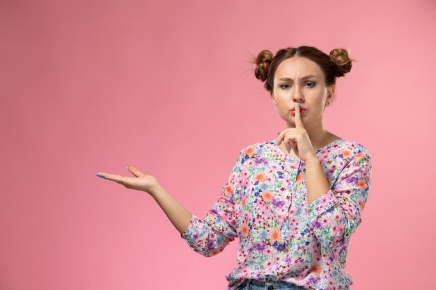 Front view young female in flower designed shirt and blue jeans showing silence sign posing on the pink background 