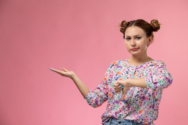 Front view young female in flower designed shirt and blue jeans showing unlike sign on the pink background 