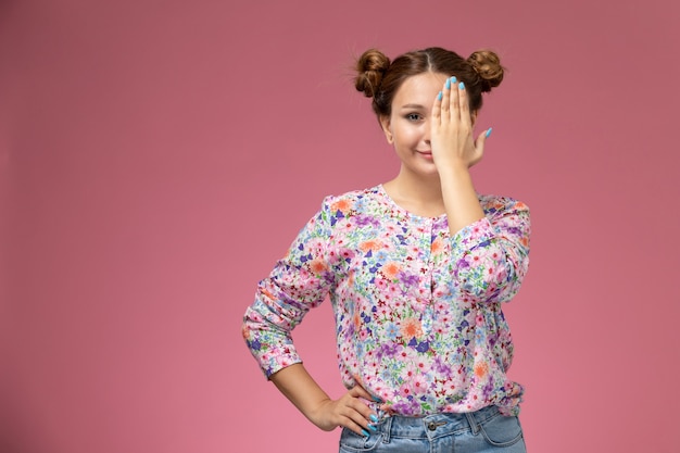 Vista frontale giovane femmina in fiore progettato camicia e blue jeans sorridente che copre un lato del viso su sfondo rosa