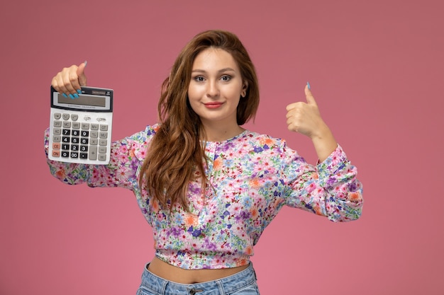 Front view young female in flower designed shirt and blue jeans smiling and holding calculator on pink background 