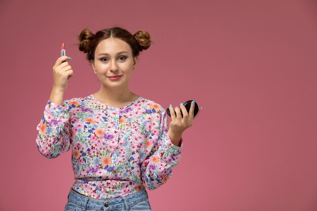Front view young female in flower designed shirt and blue jeans smiling holding lip gloss on the pink background