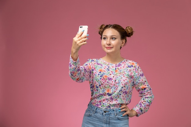 Front view young female in flower designed shirt and blue jeans taking selfie on pink background